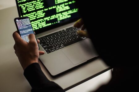 From behind crop male programmer in black hood browsing netbook and mobile phone while working in office