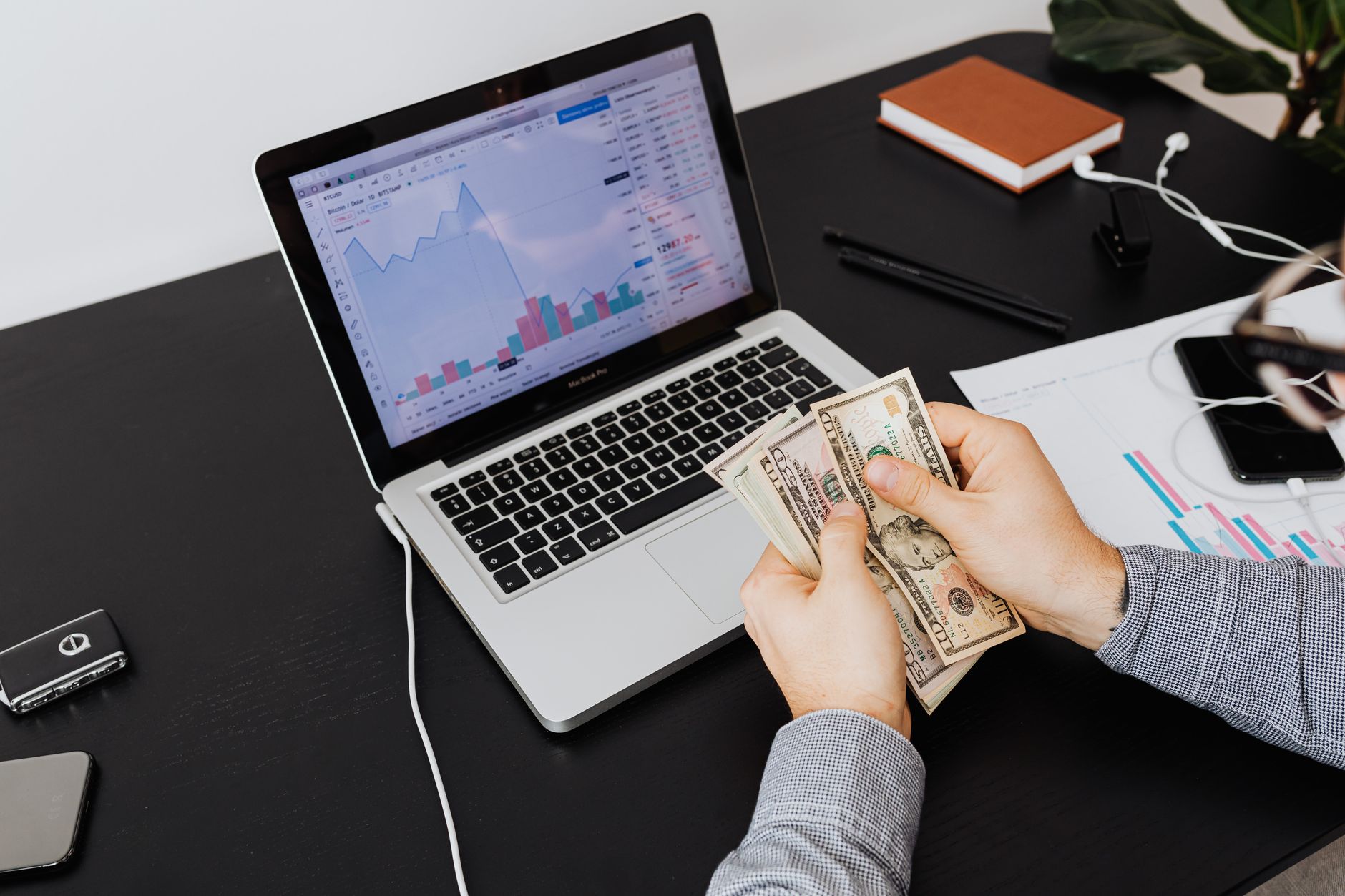 Personne comptant des billets de dollars avec des graphiques financiers sur un écran d'ordinateur portable, symbolisant l'analyse et la planification financières.