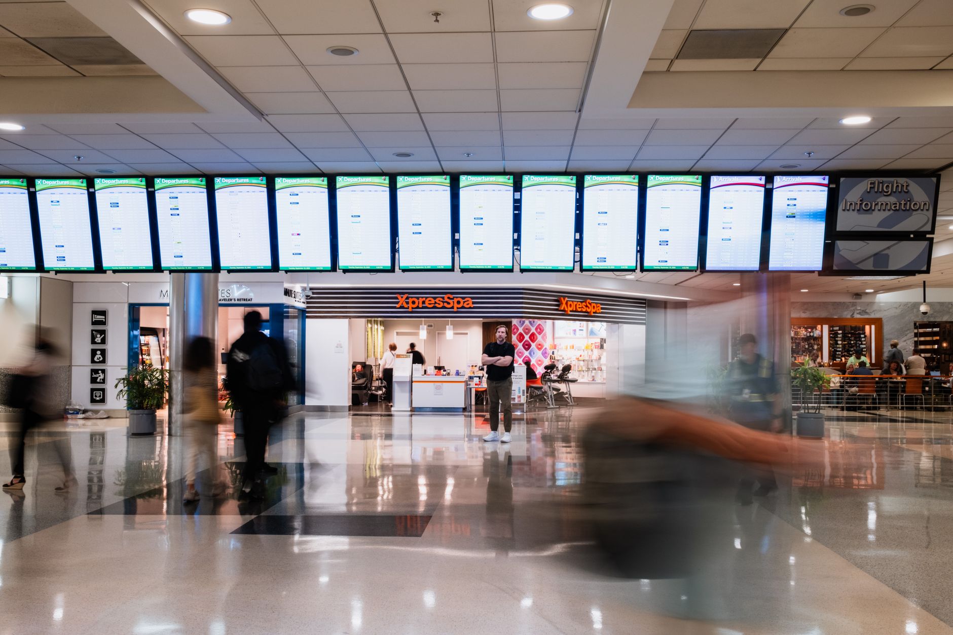 Busy travelers pass by an XpressSpa in Atlanta airport, under rows of flight information screens.