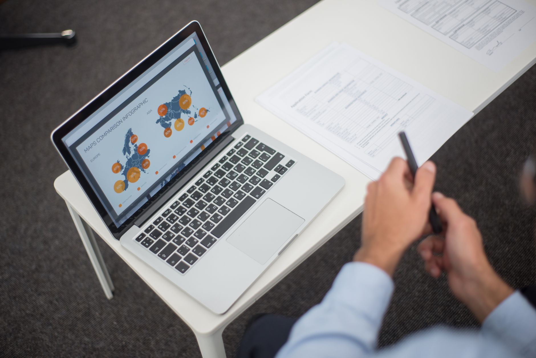 Personne en chemise bleue tenant un stylo devant un ordinateur portable affichant une infographie de cartes, à côté de documents imprimés sur une table blanche.