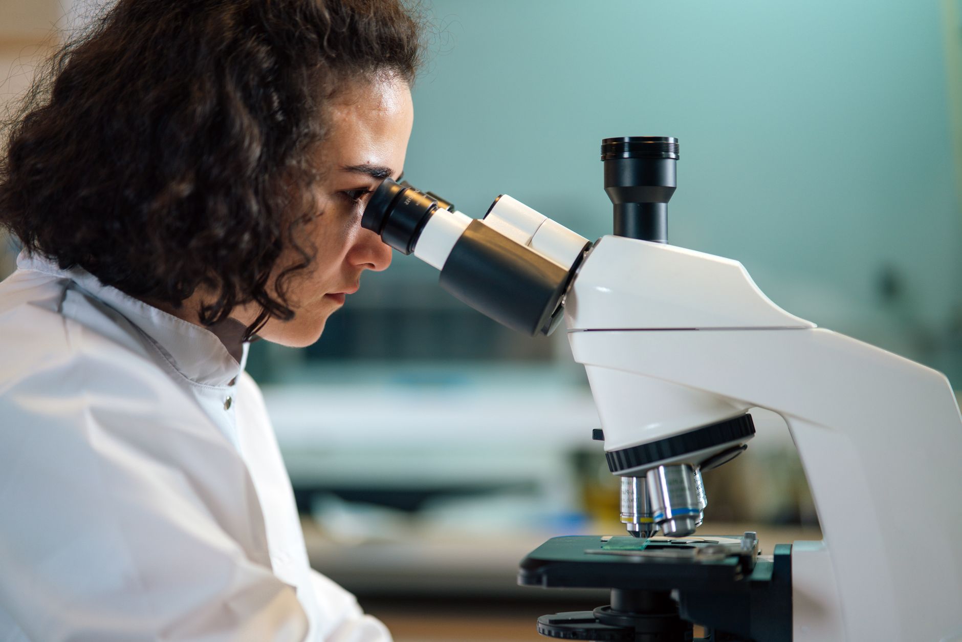 Une femme en blouse blanche regarde dans un microscope optique, dans un laboratoire dont l’arrière-plan est flou.